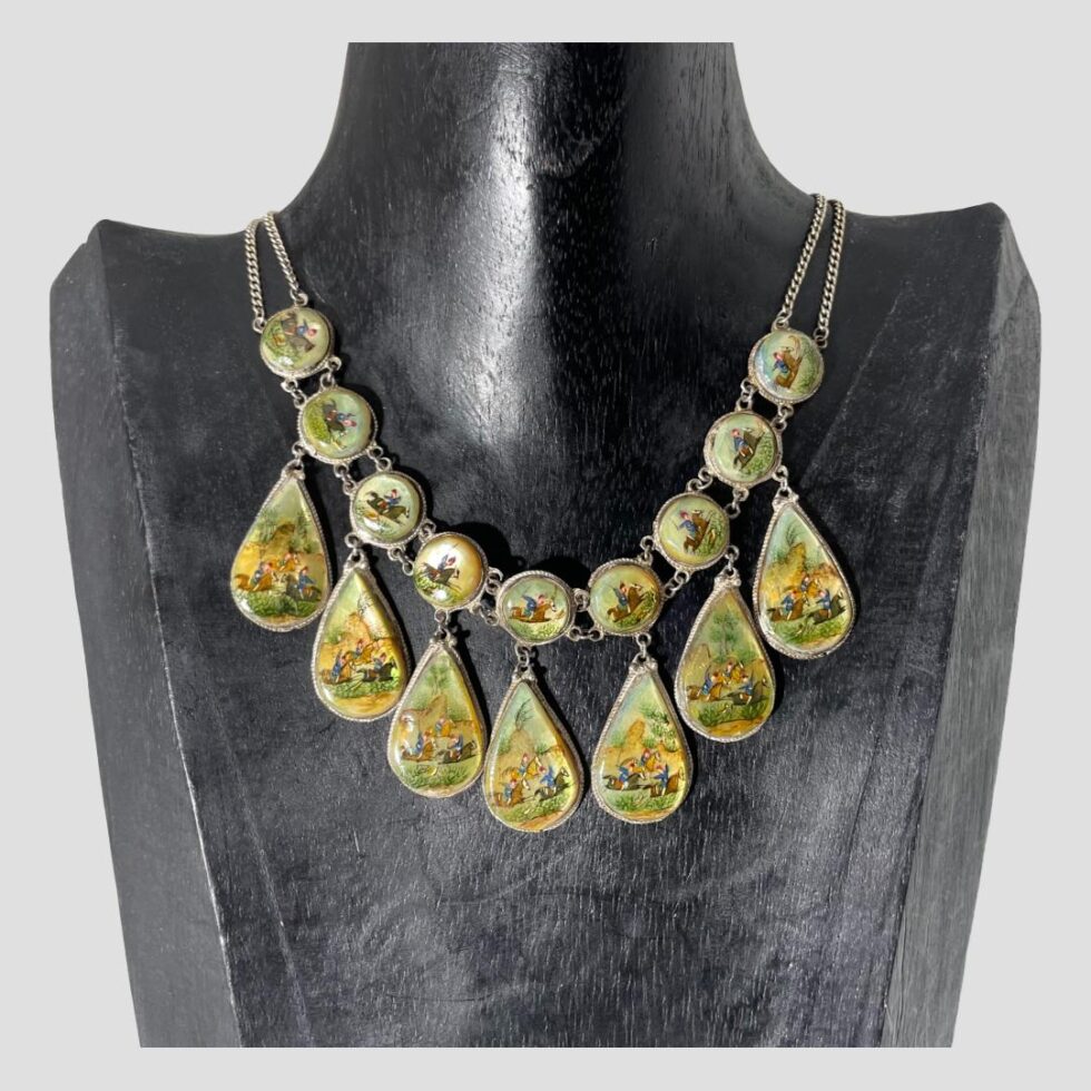 Main photo of Persian hand-painted necklace displayed on a black wooden display stand with the necklace front forward facing. The necklace consists of 9 round panels along the top with 7 teardrop shaped panels hanging below each a piece of mother of pearl. Each hand-painted scene is different showing a sequence of hunting images not dissimilar to old school cartoon making. The colours are primarily yellow and pale green with snippets of blue, white and red on the men and brown and black of the horses. The round panels have a short chain link separating with the double chain extending as the necklace chain. The teardrop panels are attached by a single ring clasp. Each panel image is incredibly detailed and beautiful.