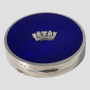 Main photo of blue guilloche enamel compact seen from a slightly raised angle looking diagonally downward to show the whole blue enamel top. The front opening side of the compact is in the foreground with the short hallmark to the left of the opener. The silver naval crown at the centre of the blue guilloche appears as if floating in the blue sea.