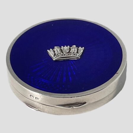 Main photo of blue guilloche enamel compact seen from a slightly raised angle looking diagonally downward to show the whole blue enamel top. The front opening side of the compact is in the foreground with the short hallmark to the left of the opener. The silver naval crown at the centre of the blue guilloche appears as if floating in the blue sea.