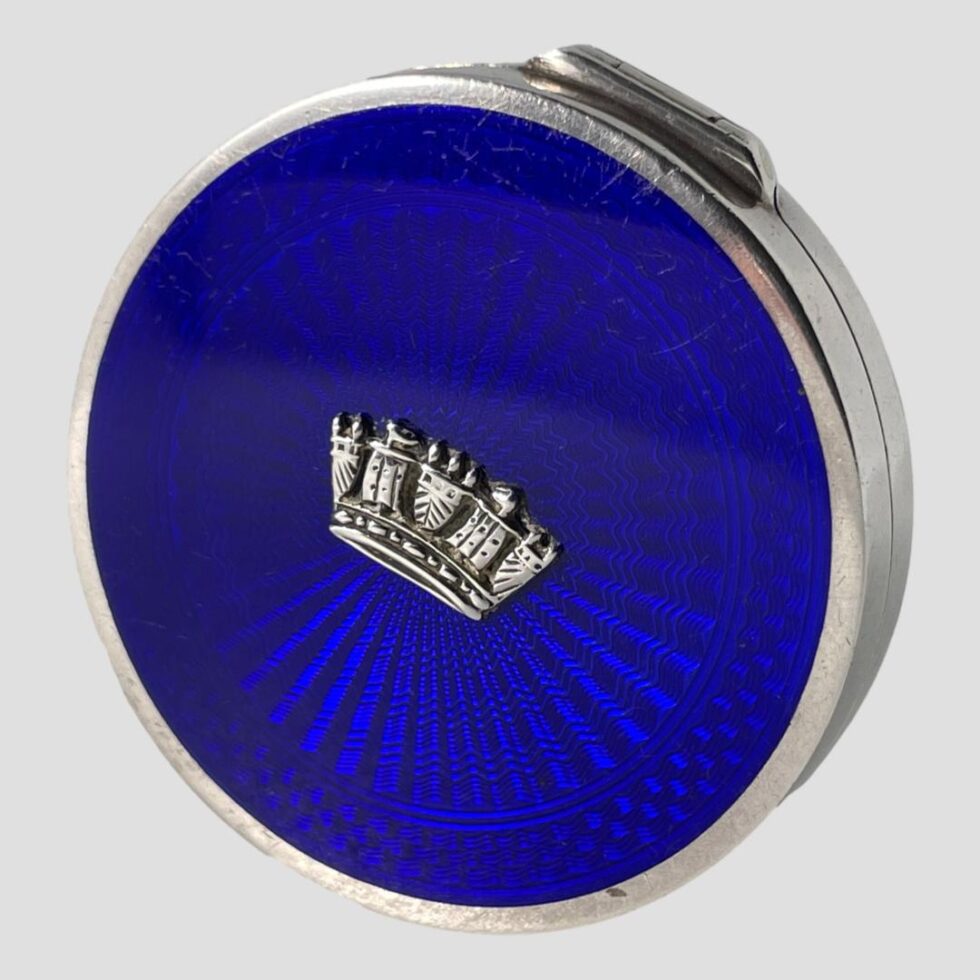 Photo of the blue guilloche enamel compact displayed on its side with the opening side face down and the hinge area visible at top. The light is reflecting on the blue enamel and creating 'ripples' in the guilloche.