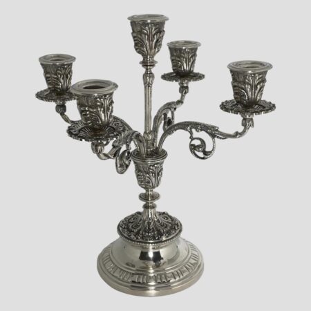 Main photo of solid Egyptian silver candelabrum displayed from an eye level with the entire piece in shot. The candelabrum has 5 sconces; tall one to the centre and 4 spread out equally on ornate arms a little lower. The candelabrum stands on a fairly plain rounded base which tapers thinner. The foot of the column has very elaborate foliage like design which is embossed. This design detail continues on the sconces and arms holding them. A sconce shaped piece sits above the base with a thinner column shooting up holding the tallest sconce. The 4 arms for the remaining 4 sconces also come out from the sconce shaped base piece giving a neat uniform appearance. Each sconce except the central one has a wax drip tray.