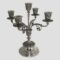 Main photo of solid Egyptian silver candelabrum displayed from an eye level with the entire piece in shot. The candelabrum has 5 sconces; tall one to the centre and 4 spread out equally on ornate arms a little lower. The candelabrum stands on a fairly plain rounded base which tapers thinner. The foot of the column has very elaborate foliage like design which is embossed. This design detail continues on the sconces and arms holding them. A sconce shaped piece sits above the base with a thinner column shooting up holding the tallest sconce. The 4 arms for the remaining 4 sconces also come out from the sconce shaped base piece giving a neat uniform appearance. Each sconce except the central one has a wax drip tray.