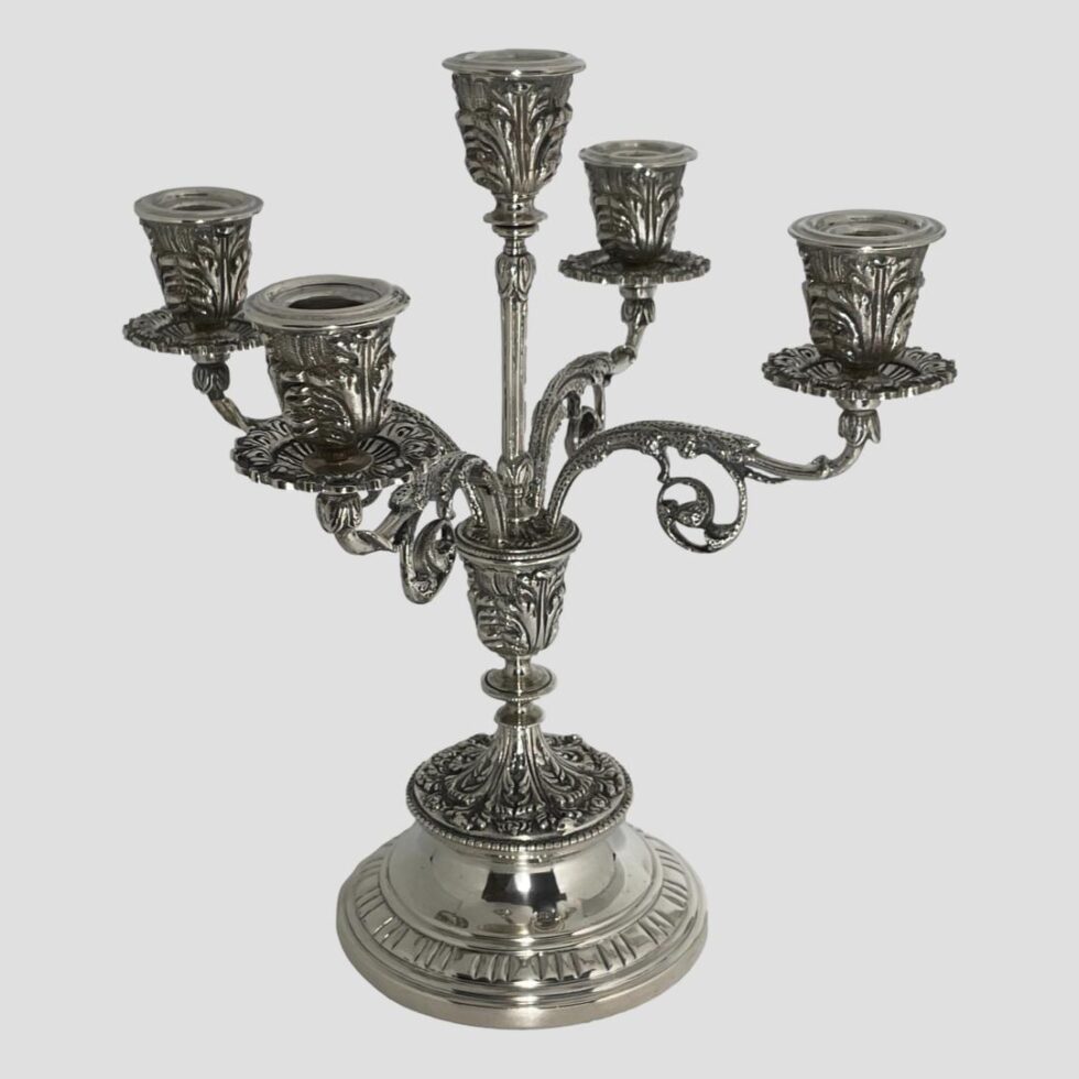 Main photo of solid Egyptian silver candelabrum displayed from an eye level with the entire piece in shot. The candelabrum has 5 sconces; tall one to the centre and 4 spread out equally on ornate arms a little lower. The candelabrum stands on a fairly plain rounded base which tapers thinner. The foot of the column has very elaborate foliage like design which is embossed. This design detail continues on the sconces and arms holding them. A sconce shaped piece sits above the base with a thinner column shooting up holding the tallest sconce. The 4 arms for the remaining 4 sconces also come out from the sconce shaped base piece giving a neat uniform appearance. Each sconce except the central one has a wax drip tray.