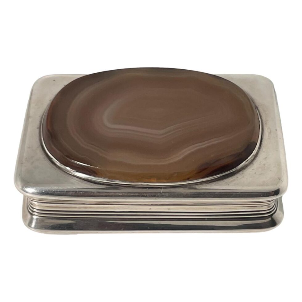 Main photo of antique silver table snuffbox seen from a raised angle looking diagonally down to the agate on the lid with parts of the surrounding silver visible all around. The box is very tactile with a smooth base and lid with rounded corners. The agate is set a little raised at the top with a silver frame surrounding it. The agate is a lovely natural colour bands in cream, pale browns and orange. The middle section of the box in slimmer than the lid and base giving a good grip to open the box lid.