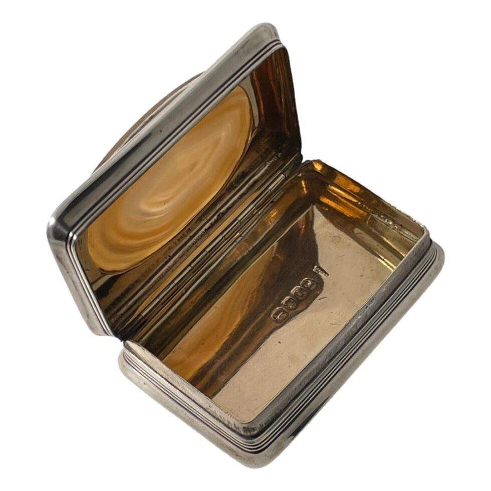 Photo looking down into an open lidded table snuffbox which is displayed diagonally across the page. The open lid is in the top left corner. The interior is gilded but has an copper colouring from the reflection of the light through the agate on the lid. The hallmark on the inside of base is just visible.