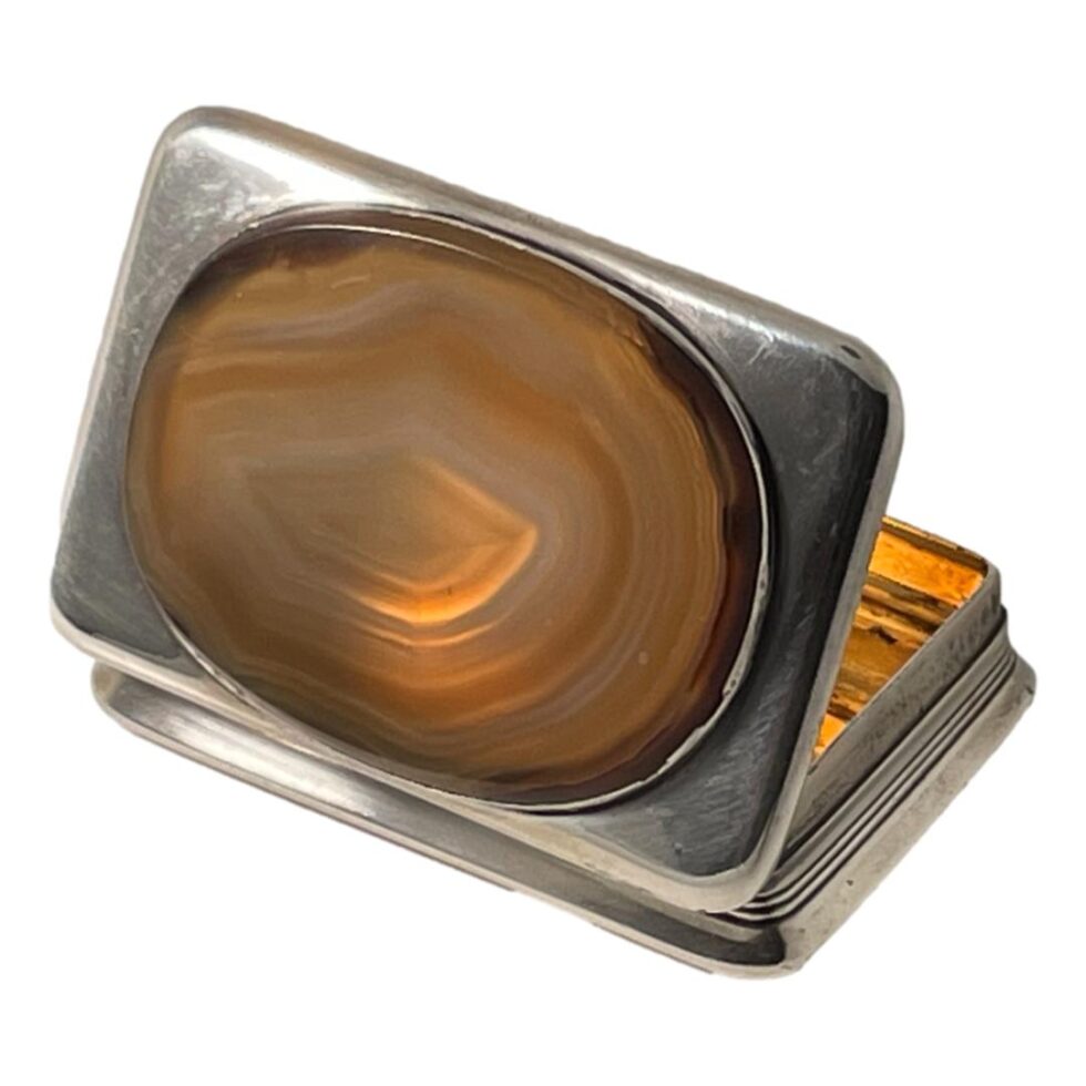 Photo of the antique silver table snuffbox seen from a back angle displayed diagonally with the lid open and in the foreground. The studio light is shining on the agate which appears to be glowing from the light and the reflection from the gilt interior. A small section of the gilt interior is visible to the right and appears glowing.