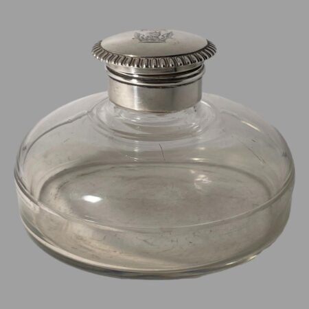 Main photo of antique silver mounted inkwell seen from an almost eye level raised very slightly to see a little of the crest on the top. The glass well has a lovely bulbous shape and flat base giving it a ships decanter type look except bulbous from the neck. The silver top is flip open with a silver neck which covers the top and inside rim.