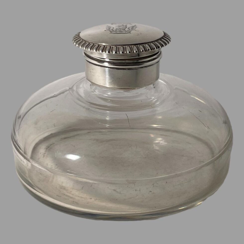 Main photo of antique silver mounted inkwell seen from an almost eye level raised very slightly to see a little of the crest on the top. The glass well has a lovely bulbous shape and flat base giving it a ships decanter type look except bulbous from the neck. The silver top is flip open with a silver neck which covers the top and inside rim.