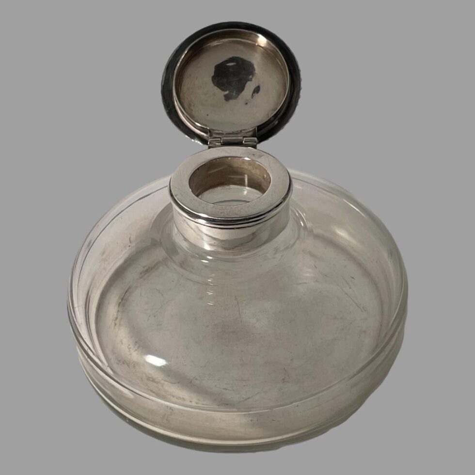 Photo of silver mounted inkwell displayed with the top fully open and to the top centre back. The inside of the lid has a little dark staining. The silver rim is visible around the mouth.