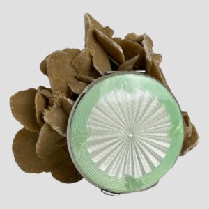Main photo of guilloche enamel mirror compact displayed upright with the peppermint green and white enamel top of lid facing forward. The mirror compact is leaning against a rock of natural desert rose. The enamel design is mesmerizing and quite hypnotic with the centre in white with wavy lines which appear to move if you look too long. The outer area around the white guilloche is a lovely shade of peppermint green with a fern like foliage at the north south east and west marks. Very Art Deco in style and look.