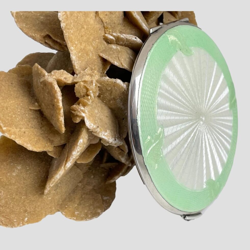 Photo of the mirror compact still upright and leaning against a rock of desert sand rose this time with the front facing towards the right showing the depth.