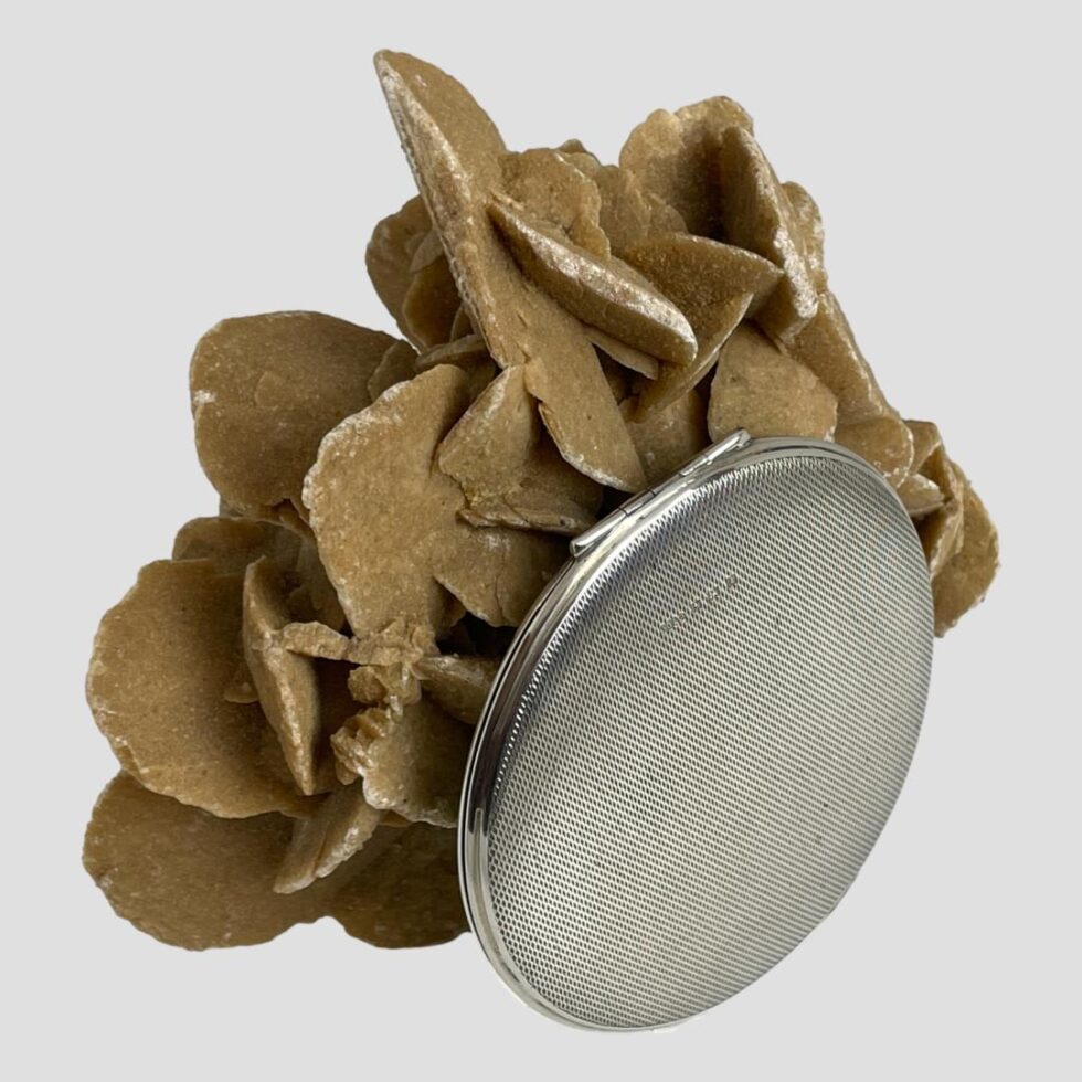 Photo of mirror compact shown upright and leaning against the rock of desert sand rose with the engine turned base on display. The hallmark is just visible almost camouflaged in the engine turned pattern of the silver.