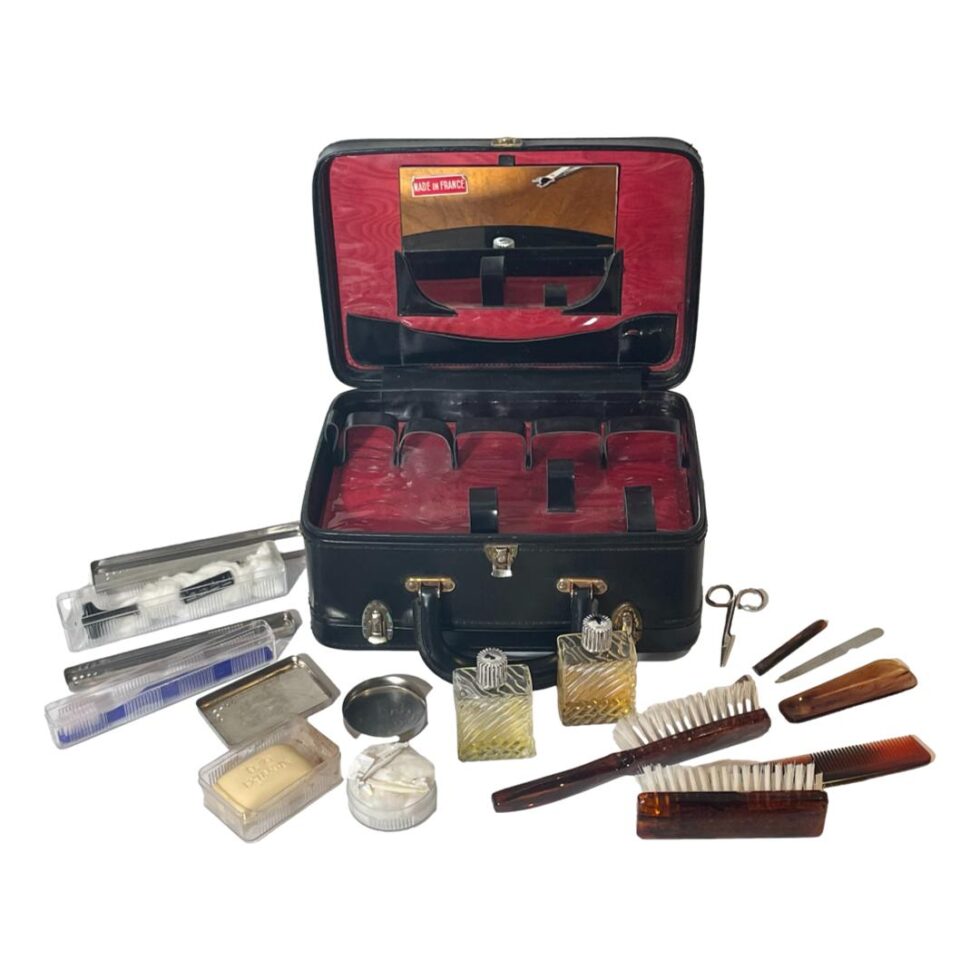 Photo of the vanity case seen from the front with lid open and all the contents laid out in front. The mirror remains inside its slot in the lid and the tray with all the fitted compartments is shown devoid of contents. Brushes and nail gear to the right, scent bottles at the centre and all the other pieces that have their own cases are to the left displayed with the lids removed and contents shown.