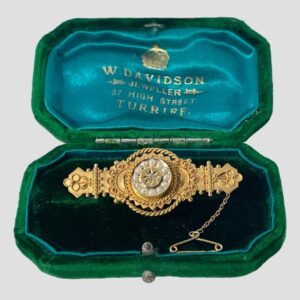 Main photo of c1906 Chester gold brooch displayed inside its original green velvet box with turquoise blue silk lining inside the lid with the Scottish retailers details in gold print. The brooch is a nice solid piece unusual for the period with a regal look about it. At the centre is a disc with diamond in the very middle inside an eight pointed star. Around this star is a frame of 12 seed pearls. The central area of the brooch is the widest with intricate gold detail including rope twists and small golden balls. This is a short and squat brooch with much intricate detail.