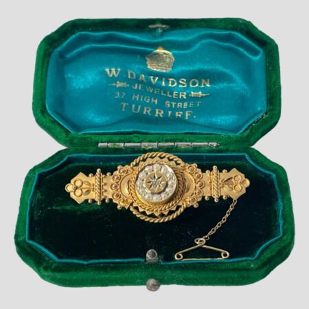 Main photo of c1906 Chester gold brooch displayed inside its original green velvet box with turquoise blue silk lining inside the lid with the Scottish retailers details in gold print. The brooch is a nice solid piece unusual for the period with a regal look about it. At the centre is a disc with diamond in the very middle inside an eight pointed star. Around this star is a frame of 12 seed pearls. The central area of the brooch is the widest with intricate gold detail including rope twists and small golden balls. This is a short and squat brooch with much intricate detail.