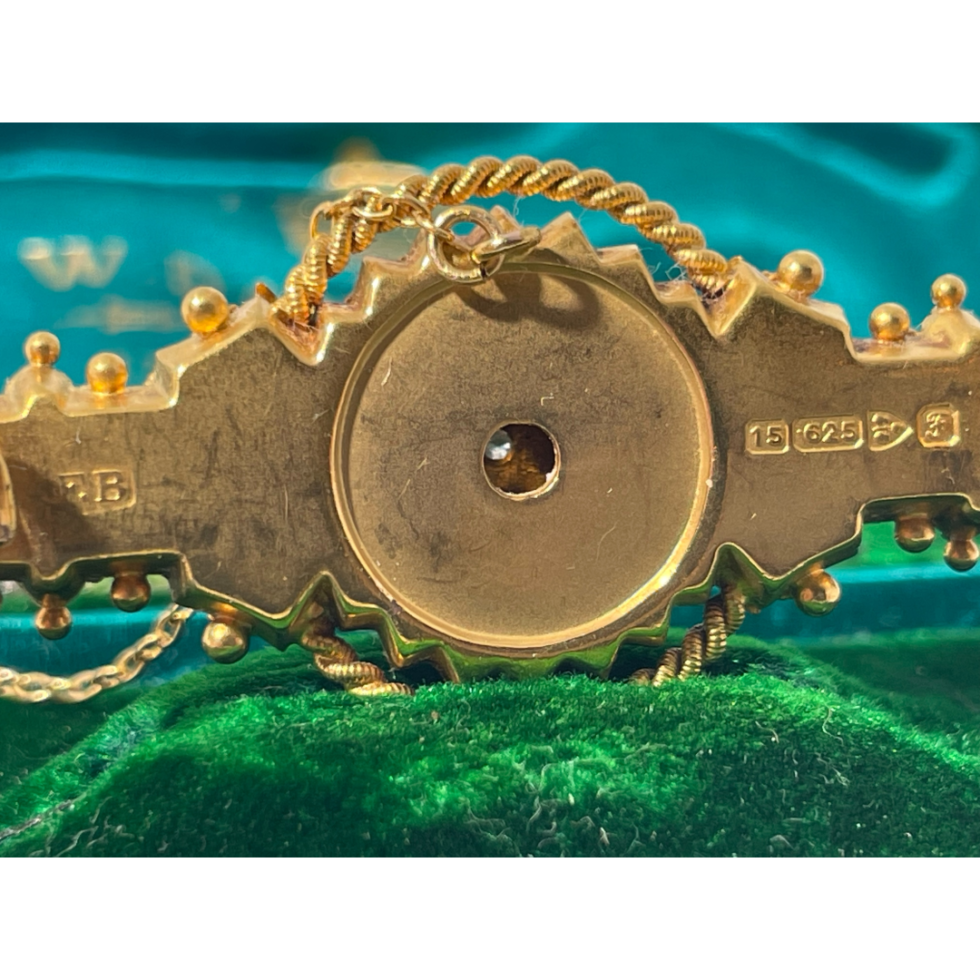 Close up of the back of brooch shown with pin open to reveal fully the full hallmark on the right and makers initials F. B on the left of the central circle.