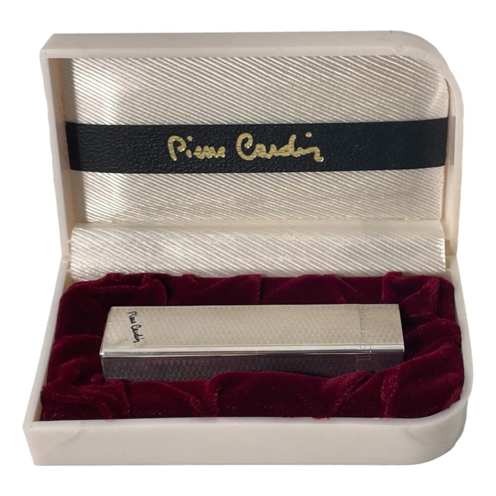 Photo of Pierre Cardin lighter displayed inside its fitted slot inside the original box. The fitted area is covered in a burgundy coloured velvet. Inside the lid is a black back horizontally across the centre with the Pierre Cardin signature across it. The Pierre Cardin signature is also on the base of the lighter in black on opposite side to the roller striker.