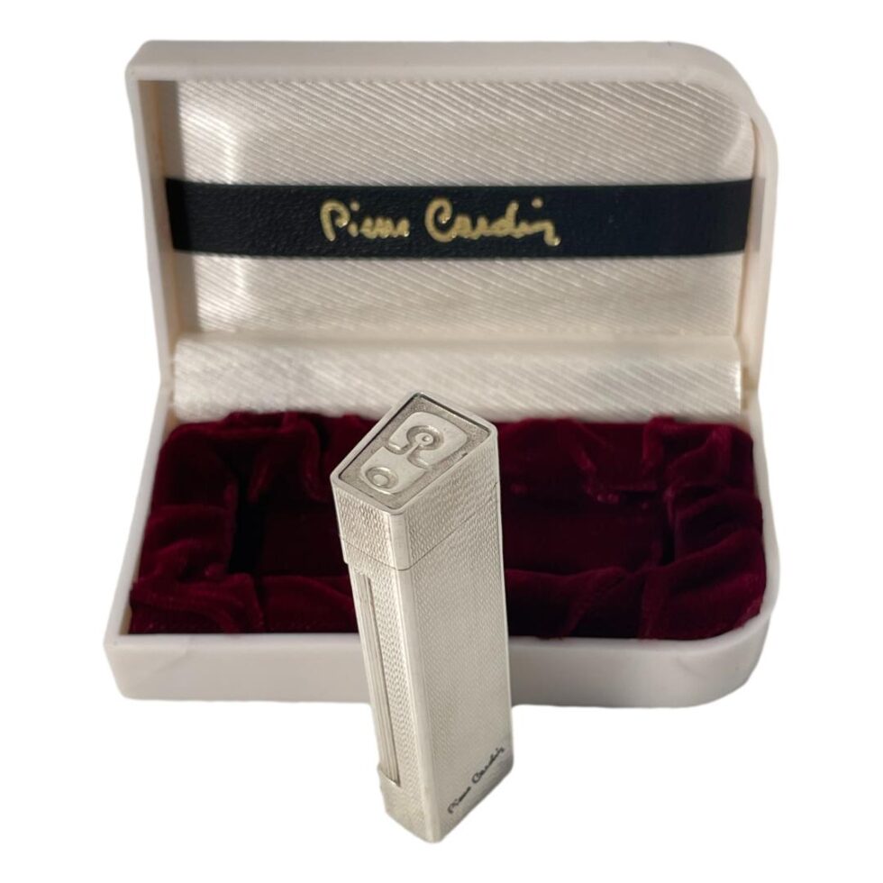 Photo of Pierre Cardin lighter displayed outside of its case and showing all the areas of logo and signature. The box is open with the lighter at the middle and we are looking down at the logo on the top of the lighter lid and the signature is visible at the bottom of the lighter.