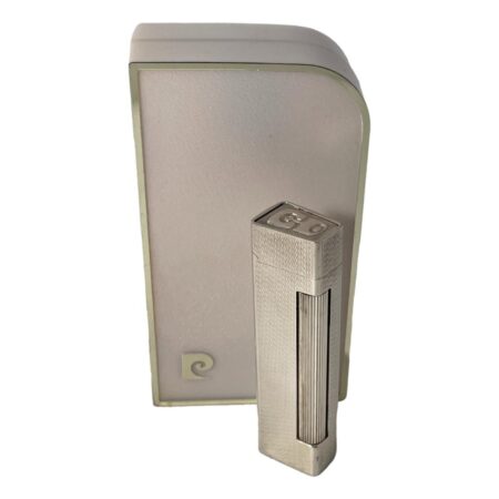 Main photo of 1970s Pierre Cardin lighter displayed outside of its original case and shown upright with the striker in the foreground. The striker for this silver coloured lighter is a long roller type that extends down almost one whole corner of the lighter. The overall design of the lighter is slimline; long & slender for easy hold. It is in good working order. The Pierre Cardin logo is on the top of the flip open lid. The original box is bigger than the lighter itself although probably perfect in relevance to size. It has a curved edge on one corner giving it a slick design of its own not to be outdone by the lighter it holds! A lovely gift item.