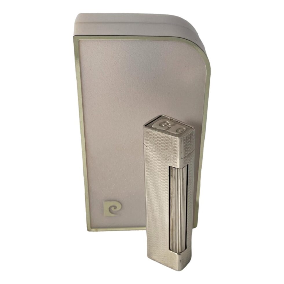 Main photo of 1970s Pierre Cardin lighter displayed outside of its original case and shown upright with the striker in the foreground. The striker for this silver coloured lighter is a long roller type that extends down almost one whole corner of the lighter. The overall design of the lighter is slimline; long & slender for easy hold. It is in good working order. The Pierre Cardin logo is on the top of the flip open lid. The original box is bigger than the lighter itself although probably perfect in relevance to size. It has a curved edge on one corner giving it a slick design of its own not to be outdone by the lighter it holds! A lovely gift item.