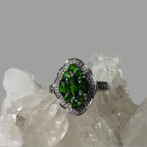 Main photo of chrome diopside silver ring displayed on top of a rock of natural quartz crystal with the ring front forward facing. The ring is oval in shape with 8 outwardly curved sides. At the centre are 3 oval cut beautiful green chrome diopside stones laid vertically with the largest at the centre. To the top and bottom of these oval stones are 3 round cut stones laid 2 down 1 up like a pyramid. A frame of cubic zirconia surrounds the chrome diopside stones. A further 6 small round cut chrome diopside stones run along the shoulders of the ring in pairs so 3 rows of 2 on each shoulder. A pretty ring with an Art Deco look.