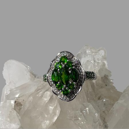 Main photo of chrome diopside silver ring displayed on top of a rock of natural quartz crystal with the ring front forward facing. The ring is oval in shape with 8 outwardly curved sides. At the centre are 3 oval cut beautiful green chrome diopside stones laid vertically with the largest at the centre. To the top and bottom of these oval stones are 3 round cut stones laid 2 down 1 up like a pyramid. A frame of cubic zirconia surrounds the chrome diopside stones. A further 6 small round cut chrome diopside stones run along the shoulders of the ring in pairs so 3 rows of 2 on each shoulder. A pretty ring with an Art Deco look.