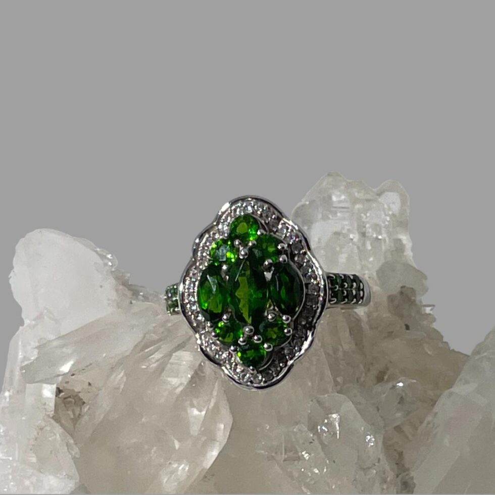 Main photo of chrome diopside silver ring displayed on top of a rock of natural quartz crystal with the ring front forward facing. The ring is oval in shape with 8 outwardly curved sides. At the centre are 3 oval cut beautiful green chrome diopside stones laid vertically with the largest at the centre. To the top and bottom of these oval stones are 3 round cut stones laid 2 down 1 up like a pyramid. A frame of cubic zirconia surrounds the chrome diopside stones. A further 6 small round cut chrome diopside stones run along the shoulders of the ring in pairs so 3 rows of 2 on each shoulder. A pretty ring with an Art Deco look.