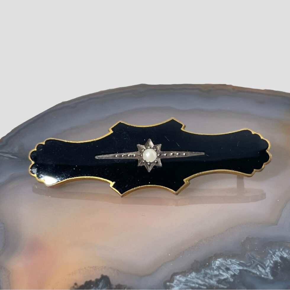Main photo of black enamel mourning brooch displayed on a slice of natural agate with the brooch front forward facing. The brooch is oblong in shape with rounded petal like ends. The centre of the brooch which holds the seed pearl is widest with blunt points as if a cross had been marked at the centre and jutted out. The seed pearl sits inside a 6 pointed star with 2 long arms to the left and right of pearl. Within these arms are 4 silver spots, the same spots hold the pearl in place at the centre. Although originally worn for mourning, this brooch would not look amiss as a regular accessory and could even work as a tie pin.