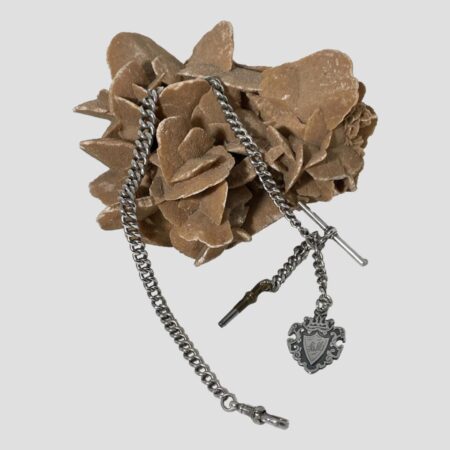 Main photo of antique silver Albert chain displayed across a rock of natural desert sand rose with the 2 ends in the foreground. On the left end is the dog clip and on right, the key winder, fob and T-bar are fanned out in the that order left to right. The chain itself is graduating with the larger links in the middle and getting smaller towards the ends.