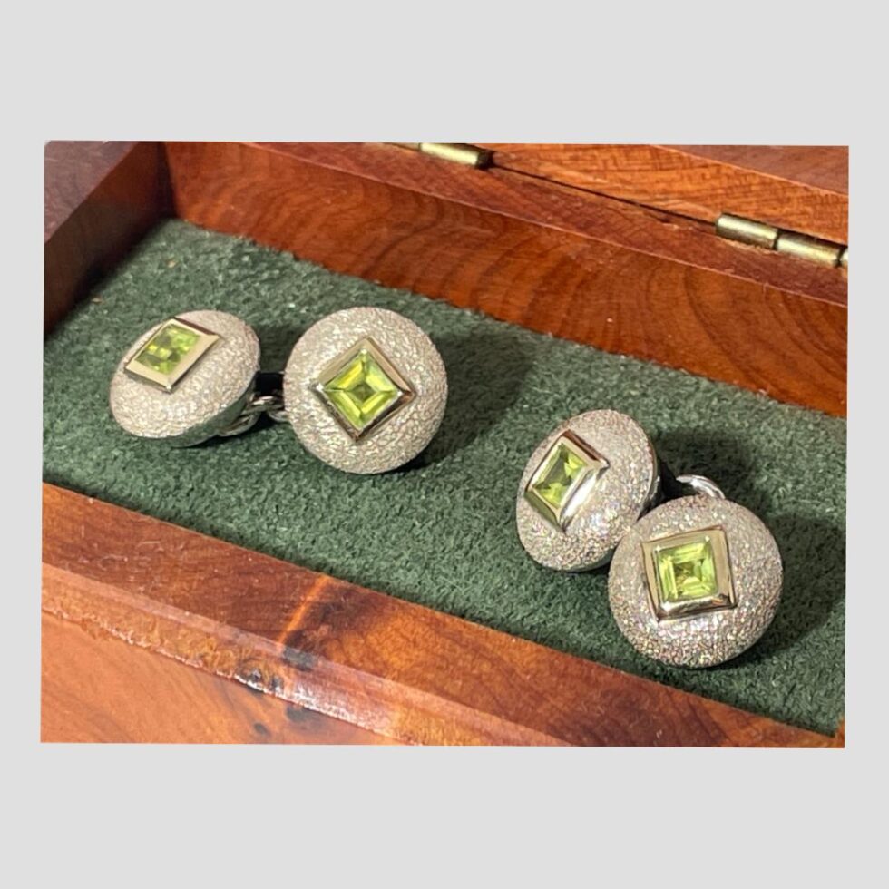 A close up of the cufflinks still inside their box and displayed with all 4 peridot sides on display. The silver discs are dome fronted and have textured effect making them look glittery. At the centre of the discs is a single square cut peridot which is inside its own gilt silver frame. This combination of colours, shapes and textures give an eye catching effect.