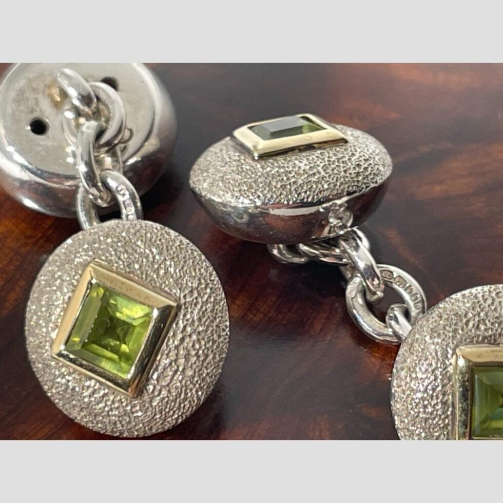 A close up the cufflinks now displayed on top of their closed wooden box. The cufflink on the left has the peridot front end in the foreground with the back on display in the back. The full hallmark is visible on the centre chain link. The cufflink on the right has both peridot fronted sides on show (one in foreground just partially) with the full hallmark much more clearly visible on the centre chain link.