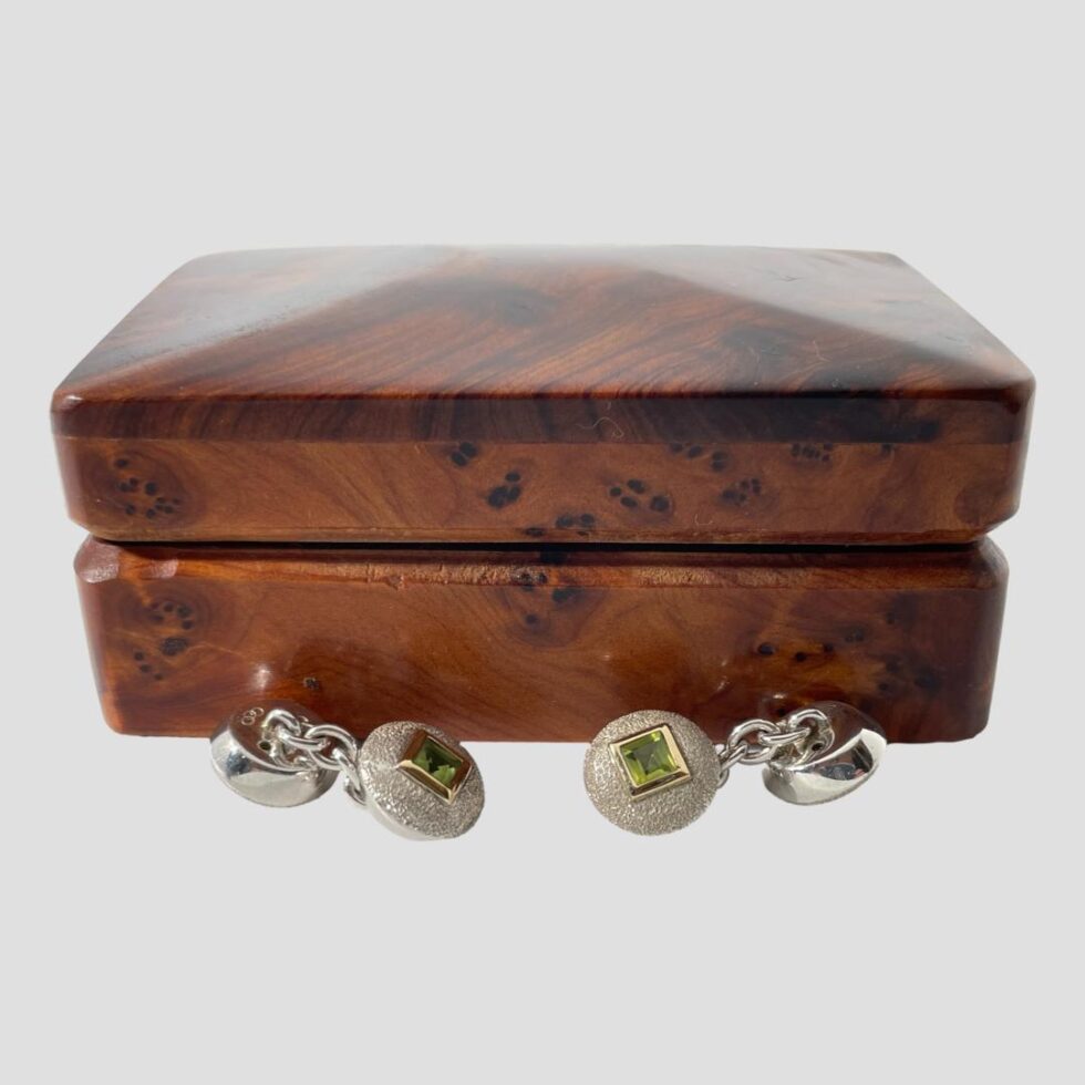 Photo of the cufflinks displayed outside of their box and placed on the surface in front. The box is shown closed. The ends at the centre are peridot side up whilst the outer ends are face down. The one on the left shows th Links of London 3 ring logo.