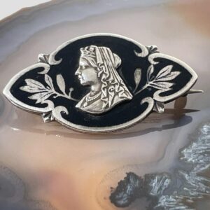 Main photo of Queen Victoria memoriam brooch displayed on top of a slice of natural agate with the brooch front forward facing. The brooch is a short oblong shape with pointed ends. It has a thin frame of silver and inlaid silver foliage to the extreme left and right. At the centre is Queen Victoria in profile in silver and this is an applied piece slightly raised from the rest of the brooch. A lovely gift item for fans of Royal memorabilia.