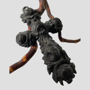 Main photo of carved bog oak cross displayed propped up against a small cherry tree twig and with the front of cross in view. The cross graduates in size being wider at the bottom. The carved floral detail extends to the whole length of the cross and has a Gothic look about it.