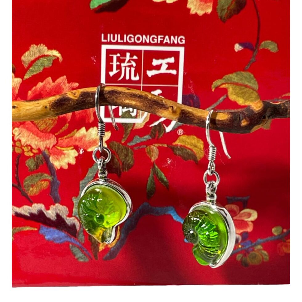 Photo of silver crystal shell earrings still hanging from the cherry tree twig this time with the lid of the box in the background. The lid and box are a very bright red with the Liuligongfang logo and name in white and decorations of flora. Even though the red is so bright, it does not actually distract from the striking green crystal.