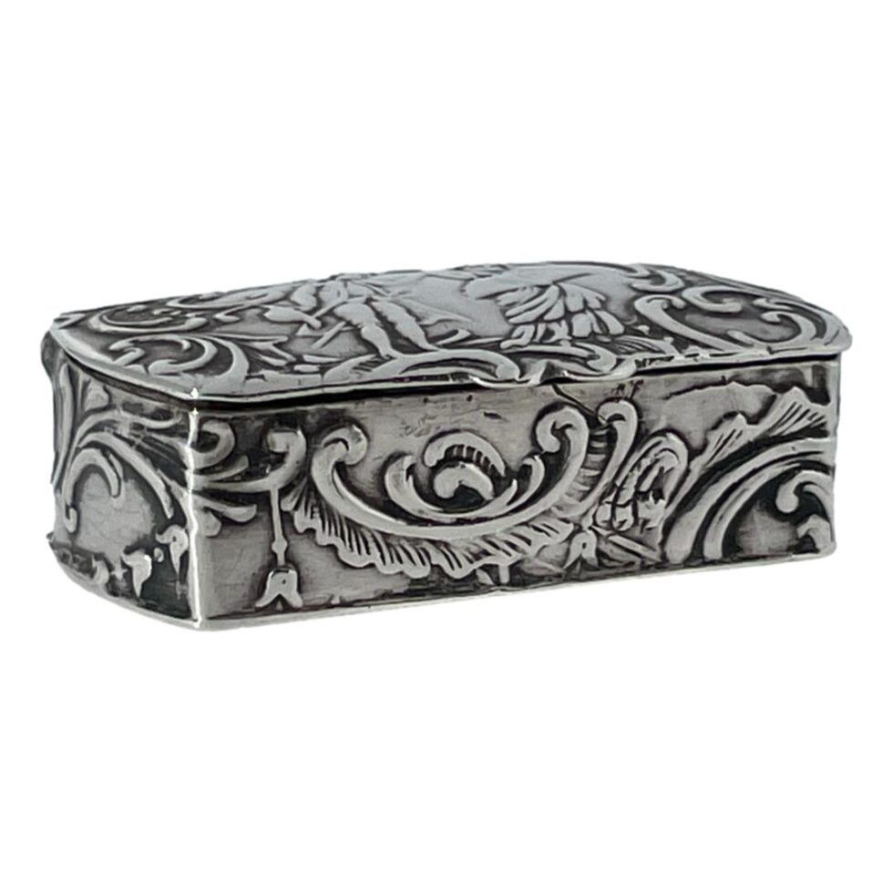 Photo of small Continental silver box seen from an eye level and displayed with one corner in the left foreground. The opening side of the box is facing towards the bottom right corner and one edge is shown on the left. The beautiful detailed foliage and scrollwork design of the box can be seen.