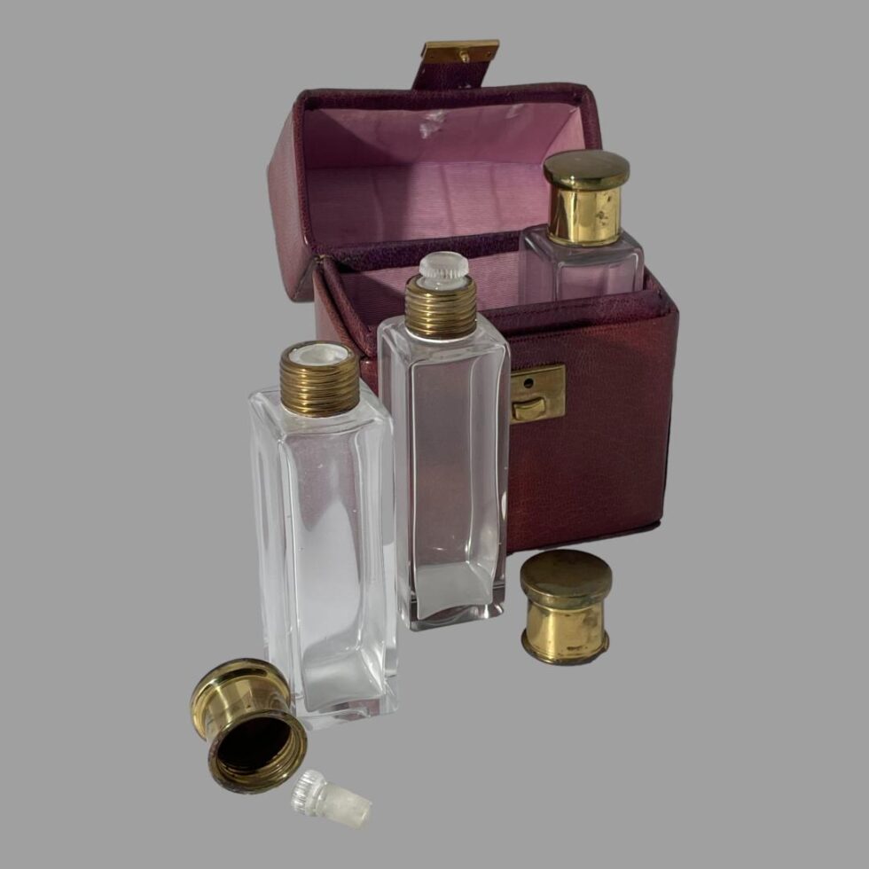 Main photo of cased travelling perfume set seen with case open. Inside the case is one bottle with brass lid in place. Just outside the case at the centre is the second bottle with brass lid removed and placed just slightly to its right and lastly, on the left is the third bottle with brass lid and glass stopper removed and placed in front of the bottle. The case looks quite purple under the studio light.