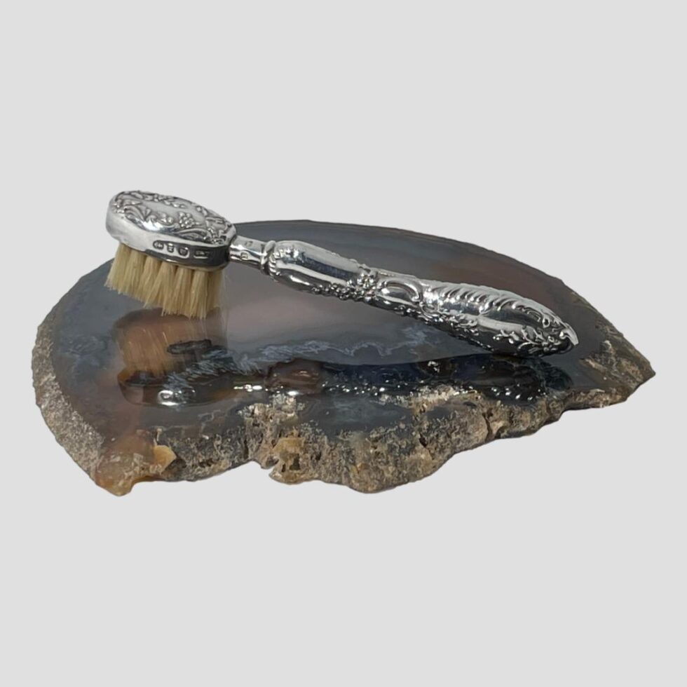 Main photo of sterling silver moustache brush displayed on top of a slice of natural agate with the brush side face down. The handle is on the right and brush head on left. The silver is beautifully and ornately embossed with floral design to back of brush and scrollwork around the handle. The full hallmark is visible around the edge of the brush head and shorter hallmark on the neck.