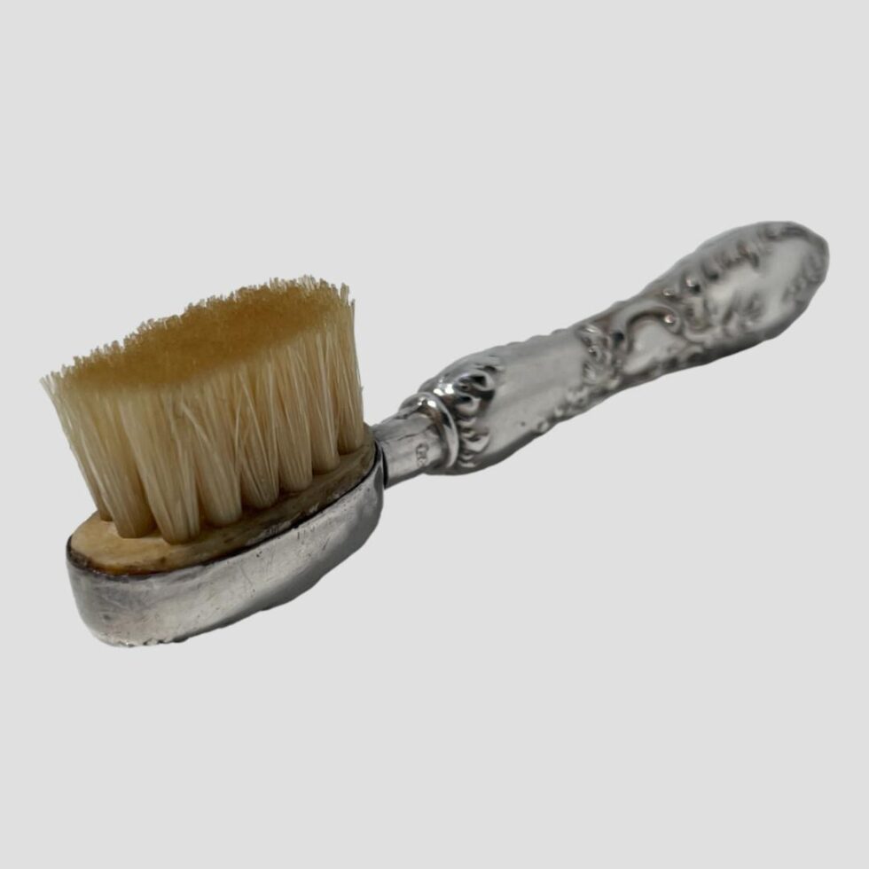 Photo of the moustache brush this time with the brush end in the bottom left corner and facing up. The bristles are mounted on wood which is encased in silver making a nice solid piece grooming kit. The brush is in very good condition for its age.