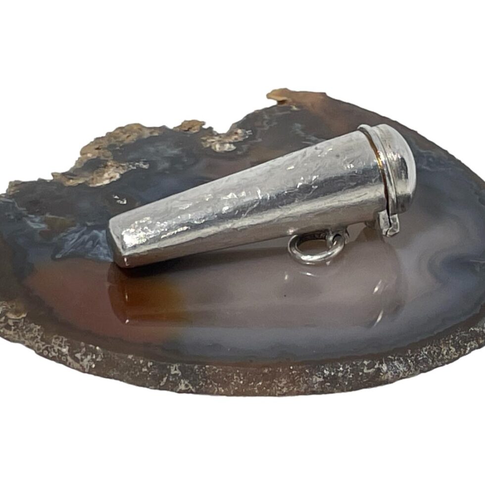 Photo of amber cheroot and case shown closed and laid on a slice of natural agate. The wider end with the lid is facing right. There is a solid ring bail just below the hinged back of lid for attaching to an Albert chain perhaps.