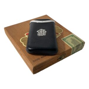 Main photo of antique leather cigar case displayed on top of an old wooden cigar box with the monogrammed side up. The case is in black leather and is a pull out style with one leather pocket fitting into an outer leather pocket. The outer pocket has the monogram and the silver edging. The monogram is very stylised an could be CFF or FFC. The leather is worn in places which is expected with the items age.
