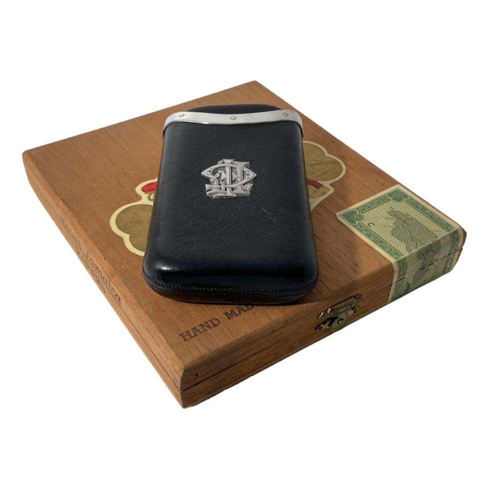 Main photo of antique leather cigar case displayed on top of an old wooden cigar box with the monogrammed side up. The case is in black leather and is a pull out style with one leather pocket fitting into an outer leather pocket. The outer pocket has the monogram and the silver edging. The monogram is very stylised an could be CFF or FFC. The leather is worn in places which is expected with the items age.