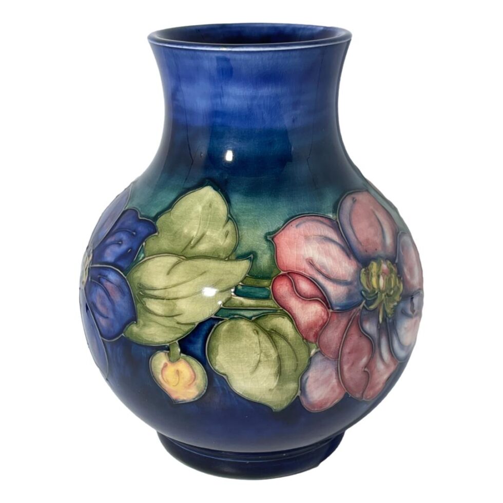 Main photo of large Moorcroft clematis vase seen from an eye level with part of a blue clematis flower on the left, pink clematis to the right and light green foliage and a yellow bud in between. The base colour of the vase is a rich blue, slightly lighter shade than the usual Moorcroft blue but beautiful all the same. Lovely bulbous shape with the floral decoration focused around the bulge!
